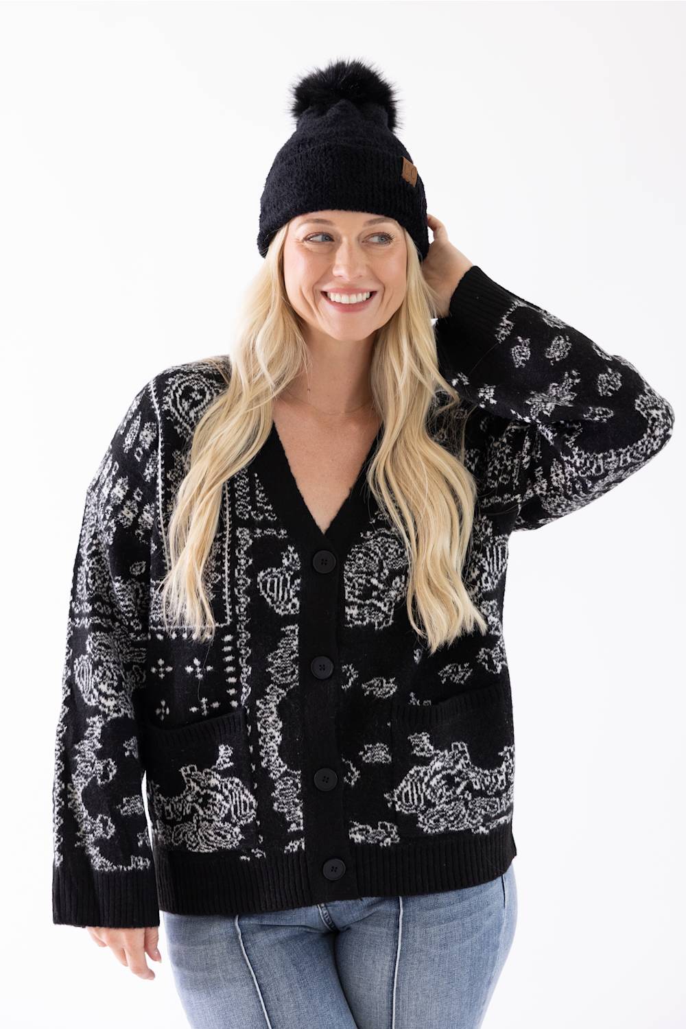 A woman smiling while wearing a black plush pom-pom beanie featuring a cuffed hem with a C.C. branded leather patch, paired with a black and white patterned cardigan.