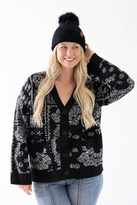 A woman smiling while wearing a black plush pom-pom beanie featuring a cuffed hem with a C.C. branded leather patch, paired with a black and white patterned cardigan.