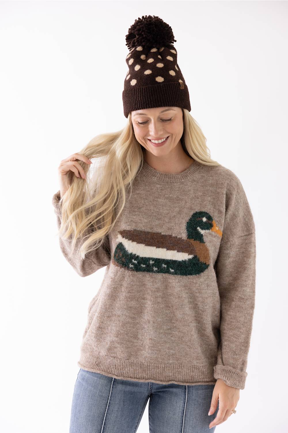 Model wearing a brown and cream polka dot beanie with a fuzzy pom-pom and a cozy beige sweater featuring a mallard duck graphic.