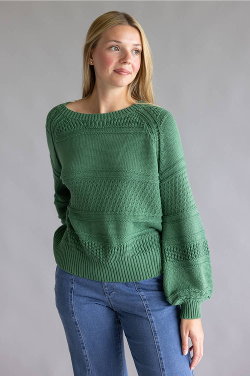 A woman modeling a green textured knit sweater with a boat neckline and blue jeans, standing against a solid gray studio background.