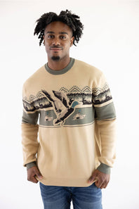 A man wearing a beige crewneck sweater with a green and black duck hunting graphic across the chest, paired with blue jeans, photographed against a plain white background.