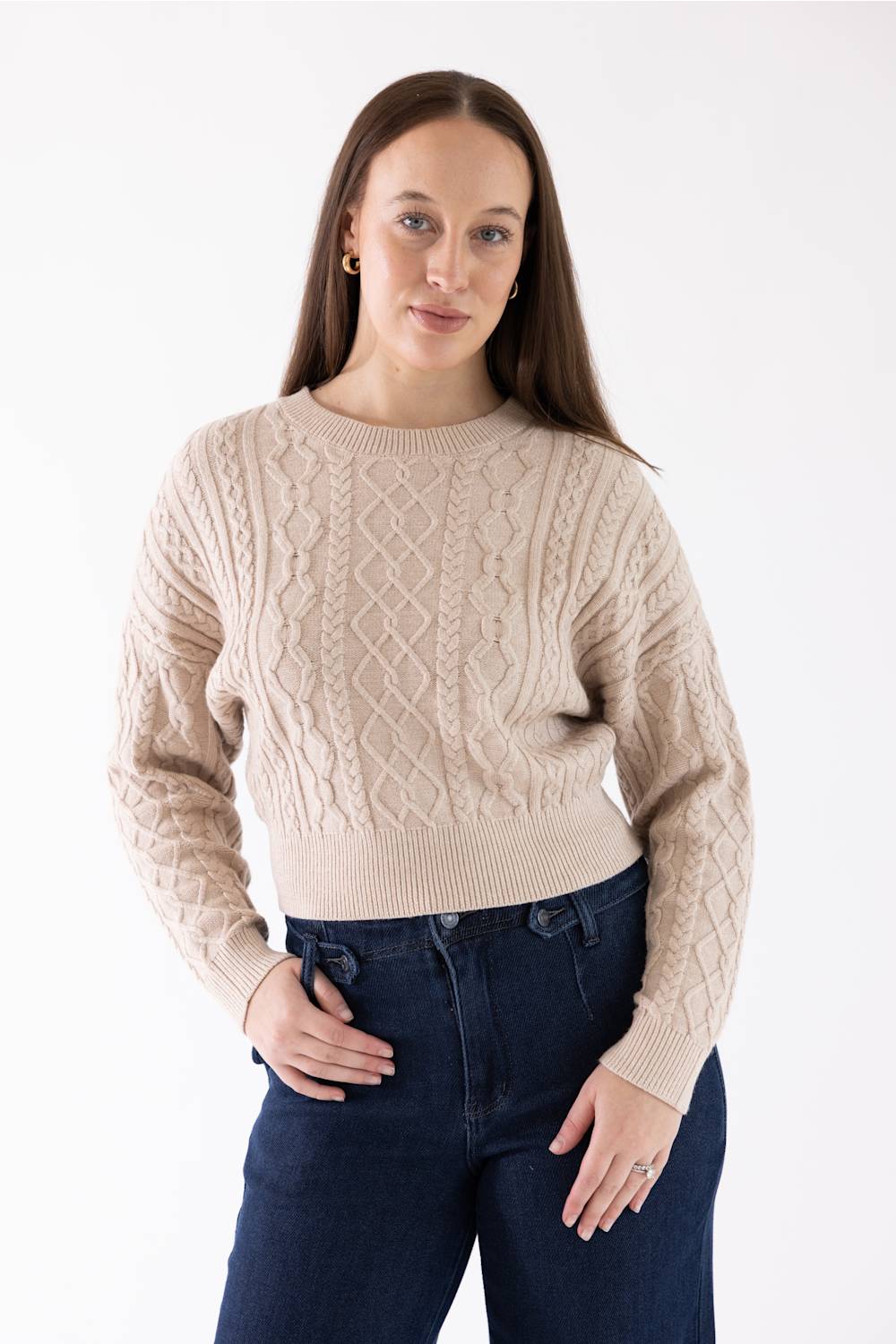 A woman wearing a beige cable-knit crewneck sweater and dark blue jeans, standing against a plain white studio background.