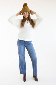 A woman modeling a white ribbed knit sweater, blue wide-leg jeans, a mustard yellow knit beanie with a pom-pom, and brown ankle boots against a plain white studio background.