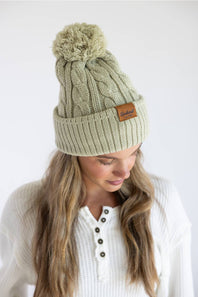 A woman wearing a light sage green cable-knit beanie with a brown pom-pom and a small brown leather brand patch on the cuff, photographed against a plain white background.