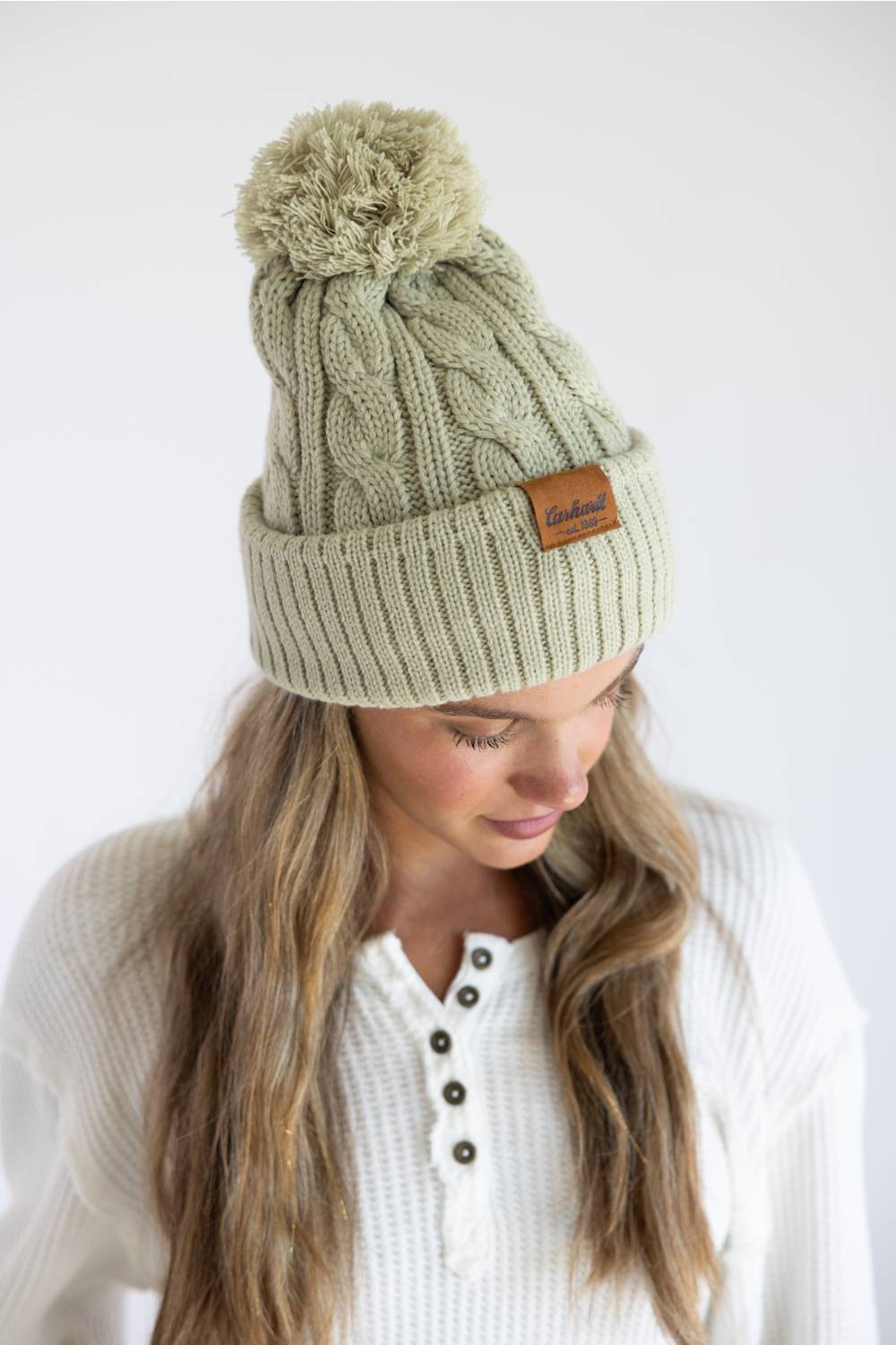 A woman wearing a light sage green cable-knit beanie with a brown pom-pom and a small brown leather brand patch on the cuff, photographed against a plain white background.