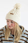 A woman wearing a cream-colored cable-knit beanie hat with a matching pom-pom and a small brown leather logo tab, paired with a blue, brown, and cream striped fleece jacket, against a plain white background.