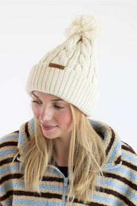 A woman wearing a cream-colored cable-knit beanie hat with a matching pom-pom and a small brown leather logo tab, paired with a blue, brown, and cream striped fleece jacket, against a plain white background.