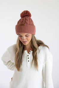 A woman wearing a dusty rose pink cable-knit beanie with a matching pom-pom and a small brown leather brand patch, paired with a white ribbed henley sweater against a plain white background.