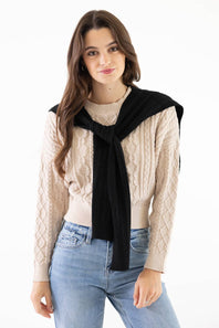 A woman modeling a beige cable-knit sweater with a black knit wrap or scarf tied around her shoulders, paired with light blue jeans, against a plain white background.