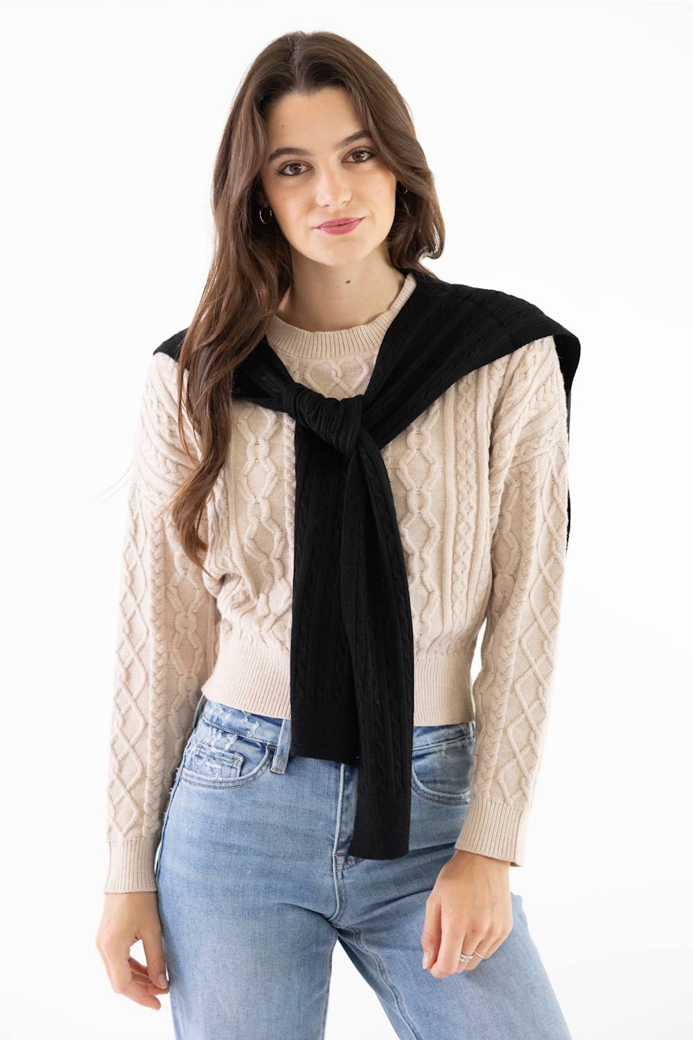 A woman modeling a beige cable-knit sweater with a black knit wrap or scarf tied around her shoulders, paired with light blue jeans, against a plain white background.