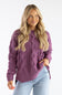 A woman with long blonde hair wearing a chunky cable-knit sweater in a dusty purple color, paired with light blue jeans, against a plain white studio background.