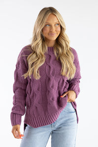 A woman with long blonde hair wearing a chunky cable-knit sweater in a dusty purple color, paired with light blue jeans, against a plain white studio background.