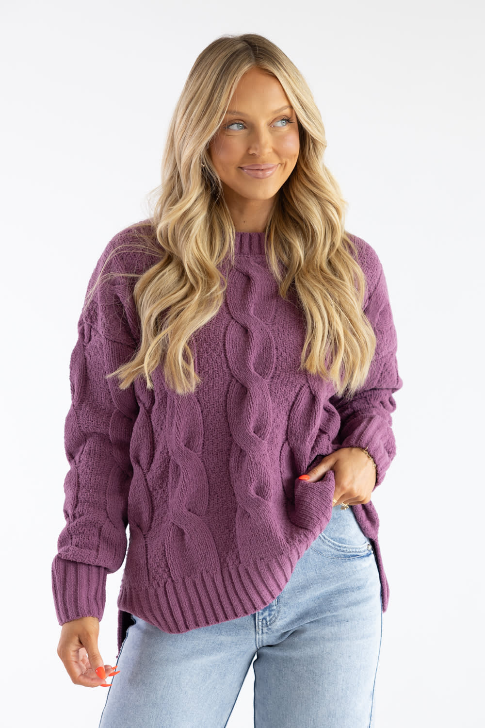 A woman with long blonde hair wearing a chunky cable-knit sweater in a dusty purple color, paired with light blue jeans, against a plain white studio background.
