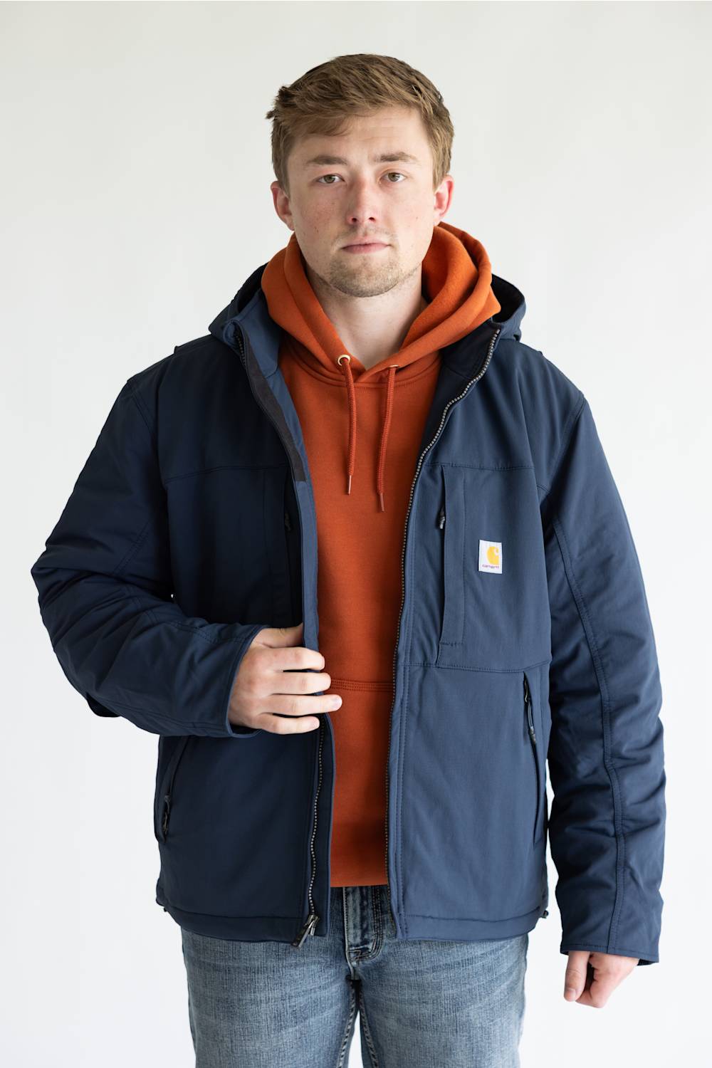 A man wearing a navy blue Carhartt jacket over an orange hoodie and blue jeans, standing against a plain light gray background.