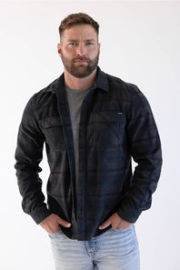 A man wearing a dark navy and black plaid button-up flannel shirt over a gray t-shirt and light blue jeans, shown from the neck down against a white background.