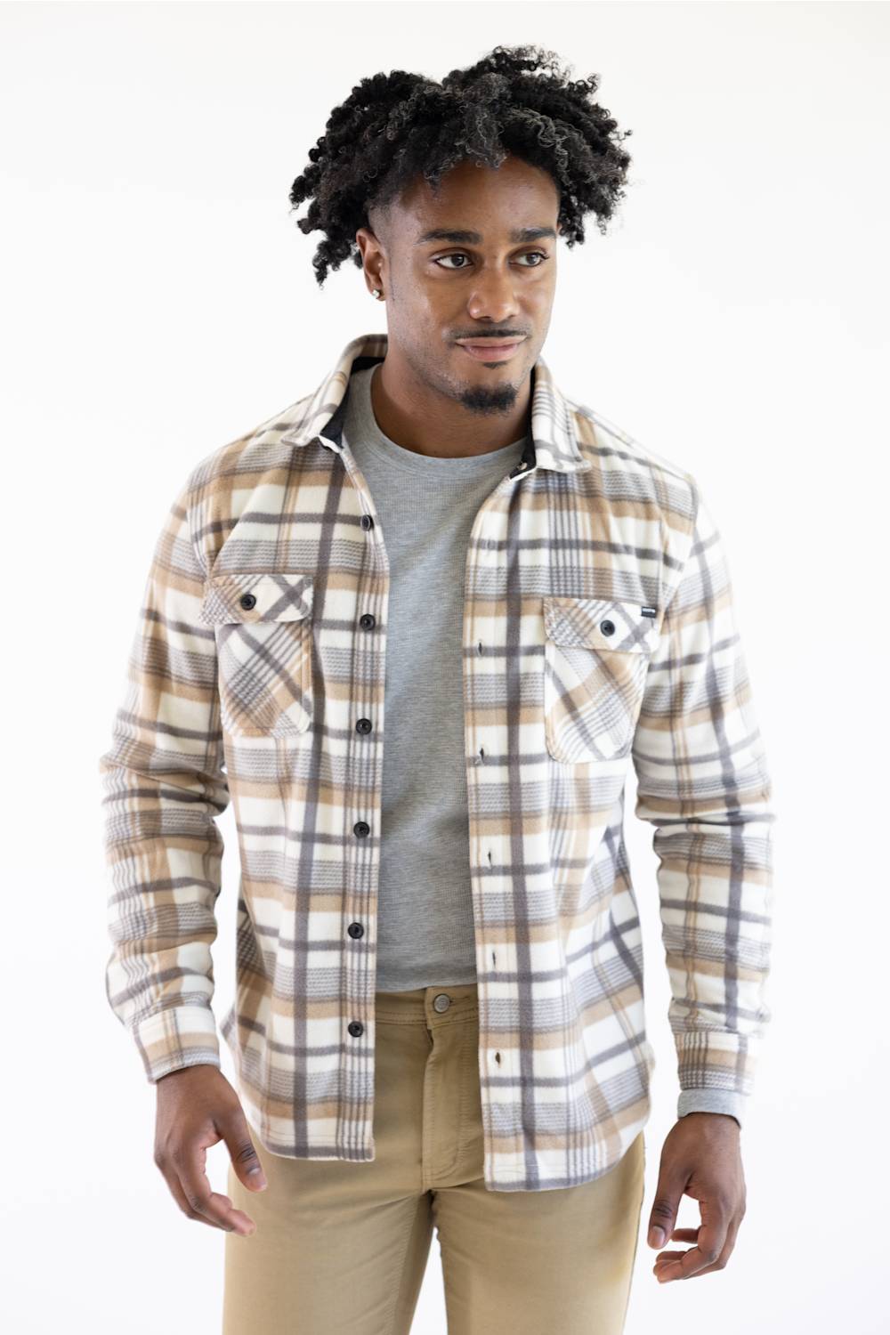 A man wearing a beige, brown, and gray plaid button-up flannel shirt over a gray t-shirt and khaki pants, photographed against a plain white studio background.