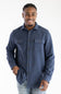 A smiling man wearing a navy blue long-sleeve button-up shirt with two chest pockets and light blue jeans, standing against a plain white studio background.