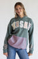 A woman wearing a grey  pullover hoodie with a large white and pink 'WRSHE' graphic across the chest, a pink and blue striped pocket, and embroidered text on the sleeves, paired with blue jeans against a white studio background.
