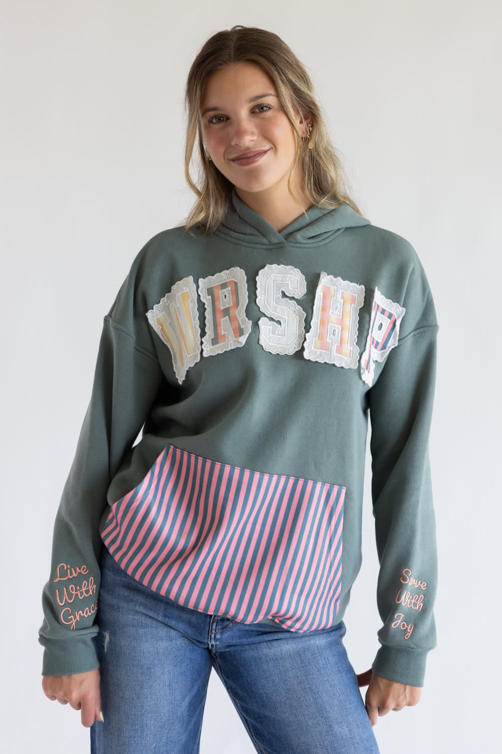 A woman wearing a grey  pullover hoodie with a large white and pink 'WRSHE' graphic across the chest, a pink and blue striped pocket, and embroidered text on the sleeves, paired with blue jeans against a white studio background.