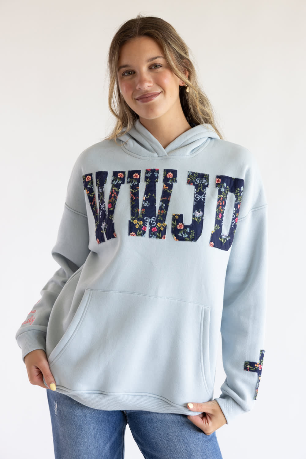 A woman wearing a light blue hoodie with a floral-patterned 'KWJD' graphic across the chest, paired with blue jeans, against a plain white background.