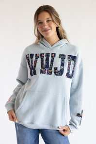 A woman wearing a light blue hoodie with a floral-patterned 'KWJD' graphic across the chest, paired with blue jeans, against a plain white background.