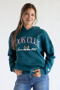 A woman wearing a teal green hoodie with 'BOOK CLUB Since 1989' printed in pink and white on the front, paired with blue jeans, against a plain white background.