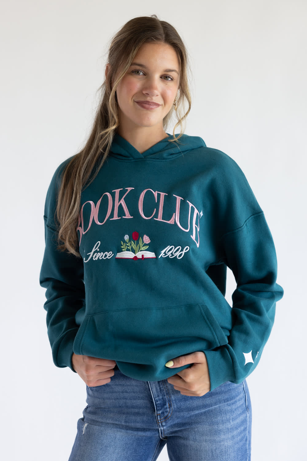 A woman wearing a teal green hoodie with 'BOOK CLUB Since 1989' printed in pink and white on the front, paired with blue jeans, against a plain white background.