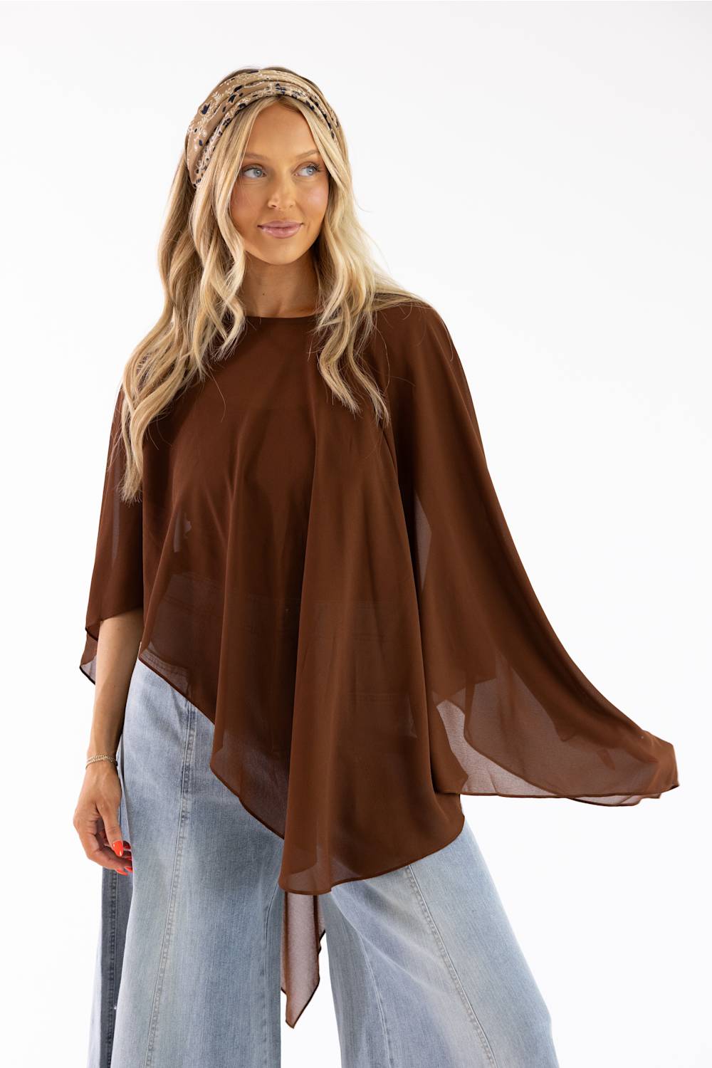 A woman modeling a brown sheer poncho-style top with wide draped sleeves and an asymmetrical hem, paired with light blue jeans and a patterned headband against a plain white background.