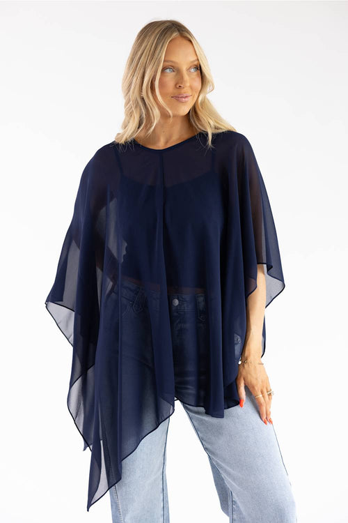 A woman modeling a sheer navy blue poncho-style top over a dark top and light blue jeans, photographed against a plain white studio background.