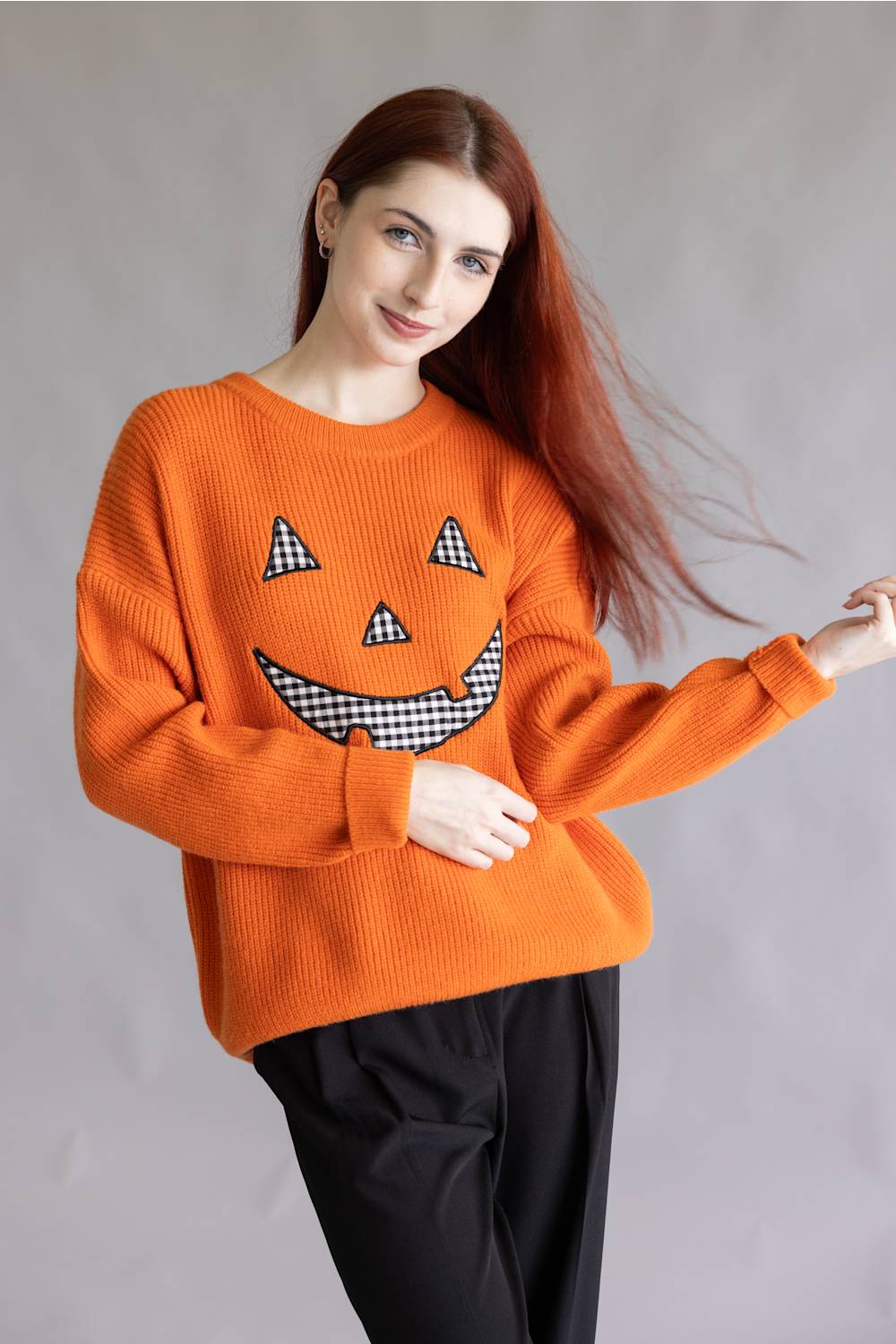 A woman with long red hair wearing a bright orange knit sweater with a black-and-white gingham jack-o'-lantern face design, paired with black pants, against a plain gray studio background.