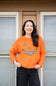A woman smiling while wearing a bright orange knit sweater with a black-and-white checkered jack-o'-lantern face on the front, standing in front of a house window.
