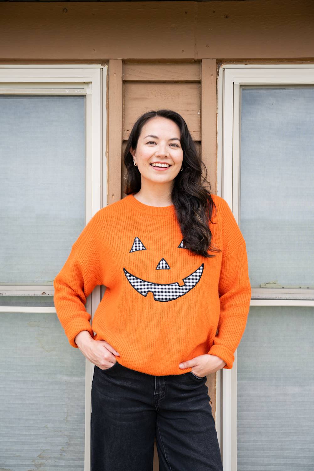 A woman smiling while wearing a bright orange knit sweater with a black-and-white checkered jack-o'-lantern face on the front, standing in front of a house window.
