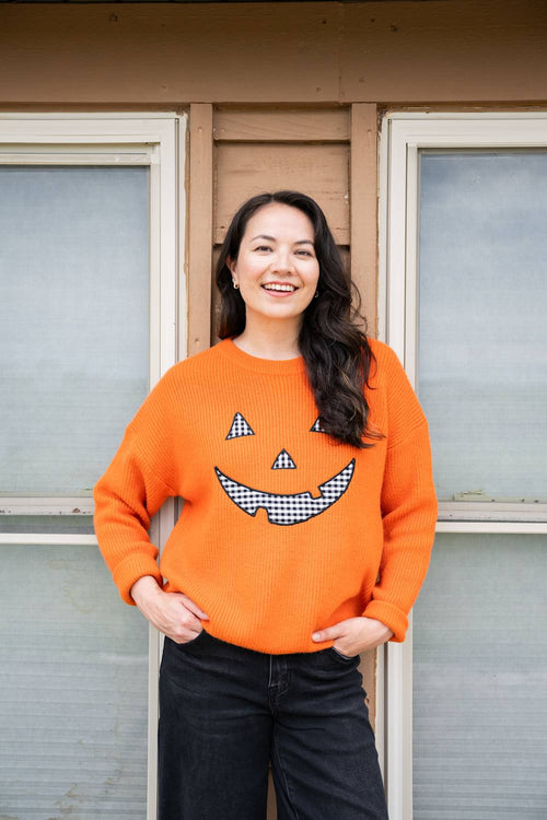 A woman smiling while wearing a bright orange knit sweater with a black-and-white checkered jack-o'-lantern face on the front, standing in front of a house window.