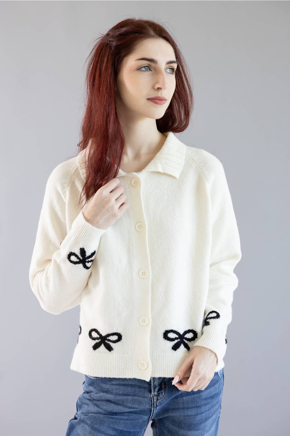 A woman with red hair wearing a cream-colored knit cardigan with black bow details on the front and cuffs, paired with blue jeans, against a solid gray studio background.