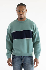A man wearing a sage green crewneck sweatshirt with a thick navy blue horizontal stripe across the chest, paired with blue jeans, against a plain white background.