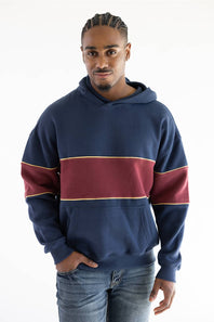 A man wearing a navy blue hoodie with a wide burgundy horizontal stripe across the chest and thin gold trim, paired with blue jeans, against a plain white background.