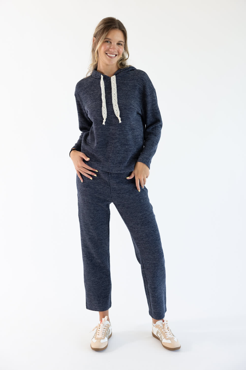 A woman modeling a matching navy blue knit loungewear set consisting of a  hoodie with cream drawstrings and matching cropped pants, paired with beige sneakers, against a plain white studio background.