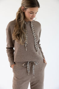 A woman wearing a matching ribbed knit lounge set in sand brown, featuring a hooded sweatshirt with leopard-print drawstrings and coordinating pants, against a plain white background.