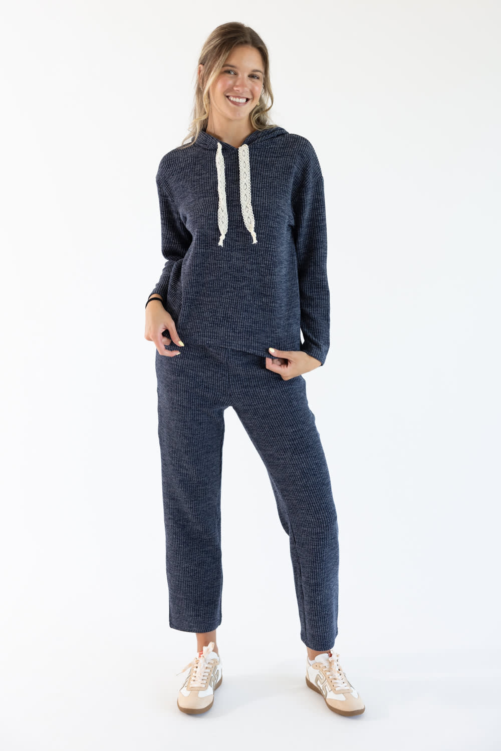 A woman modeling a matching navy blue loungewear set consisting of a long-sleeve hooded sweatshirt with white drawstrings and matching lounge pants, paired with beige sneakers, against a plain white studio background.