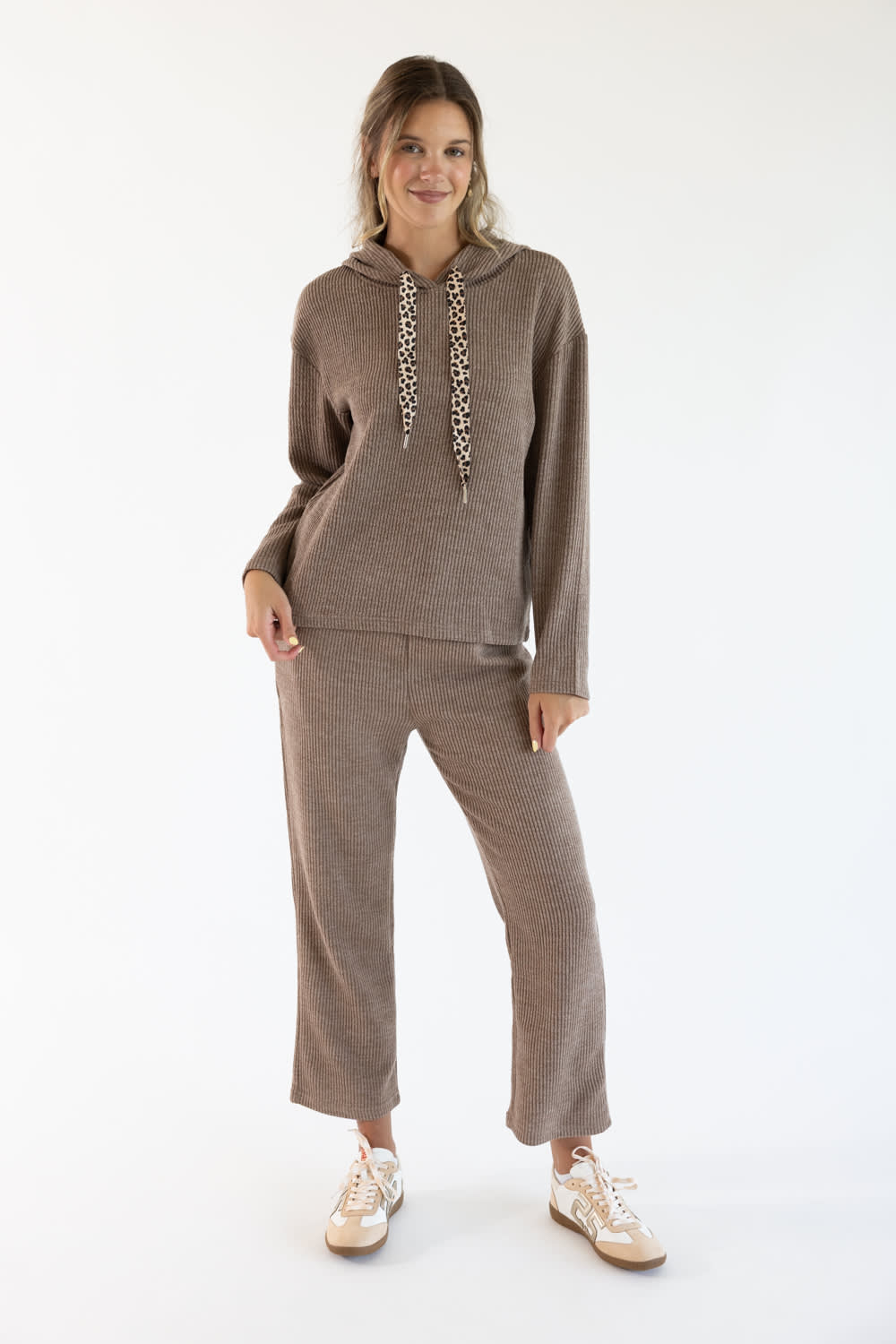 A woman modeling a matching taupe knit loungewear set consisting of a long-sleeve hoodie with patterned drawstrings and corduroy lounge pants, paired with white sneakers, against a plain white studio background.