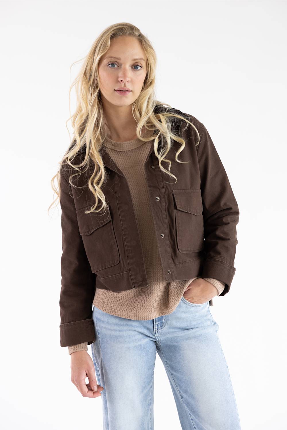 A blonde woman modeling a dark brown cropped utility jacket over a beige ribbed sweater and light blue jeans, photographed against a clean white studio background.