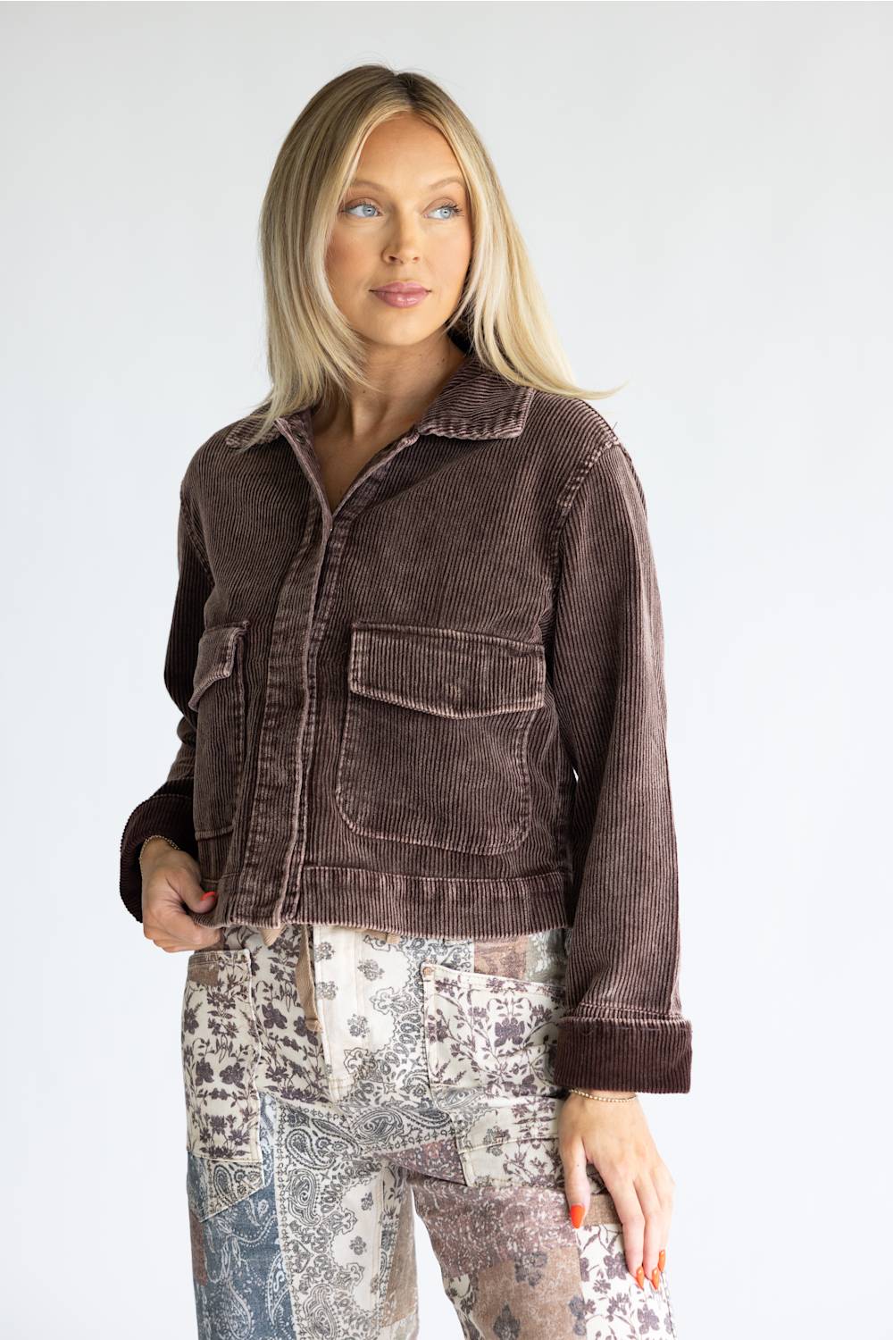 A woman modeling a dark brown corduroy jacket with front flap pockets, paired with patterned patchwork pants, against a light gray studio background.