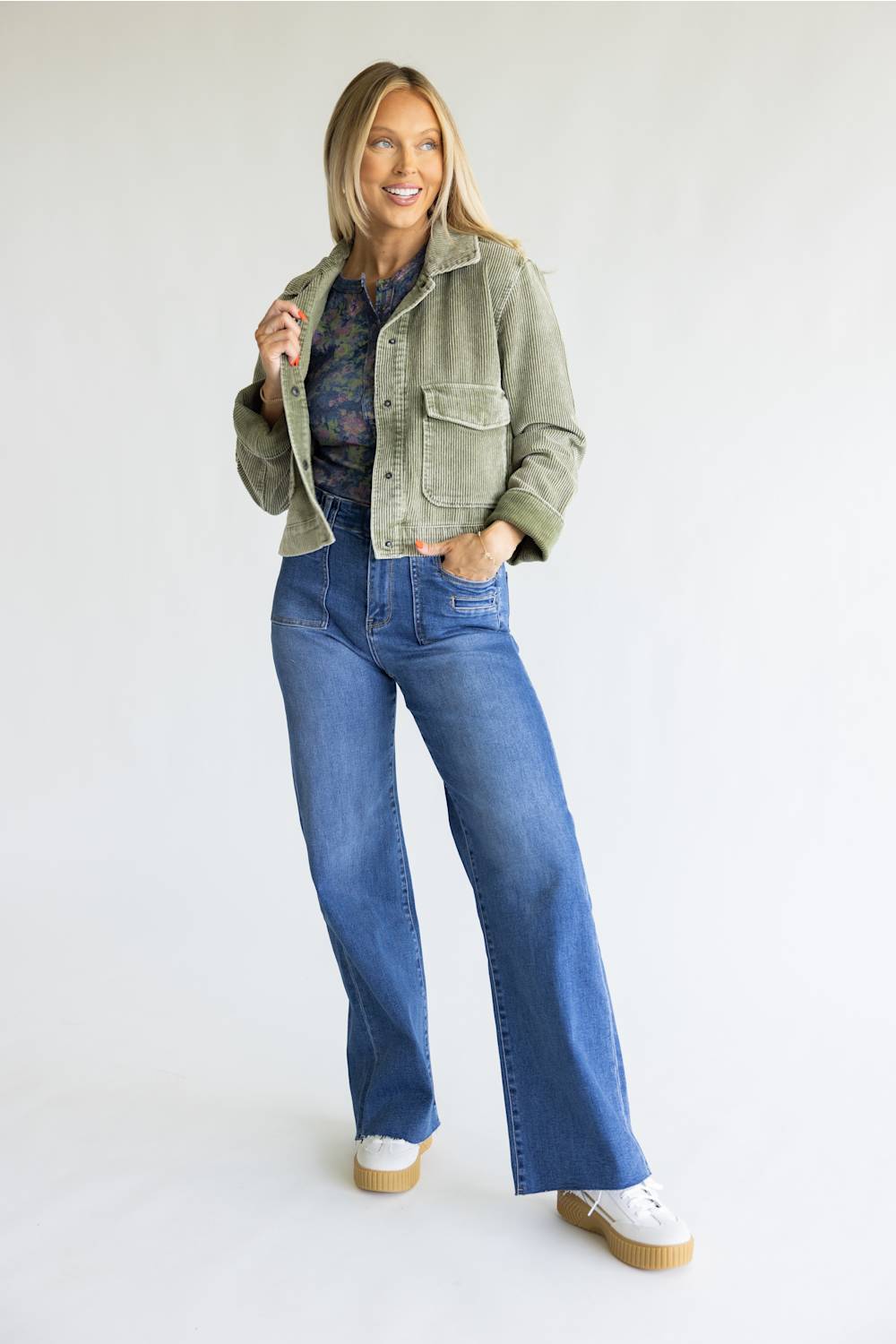 A woman modeling a pair of wide-leg blue jeans with an olive green jacket, a floral shirt, and white platform sneakers, standing against a plain white studio background.