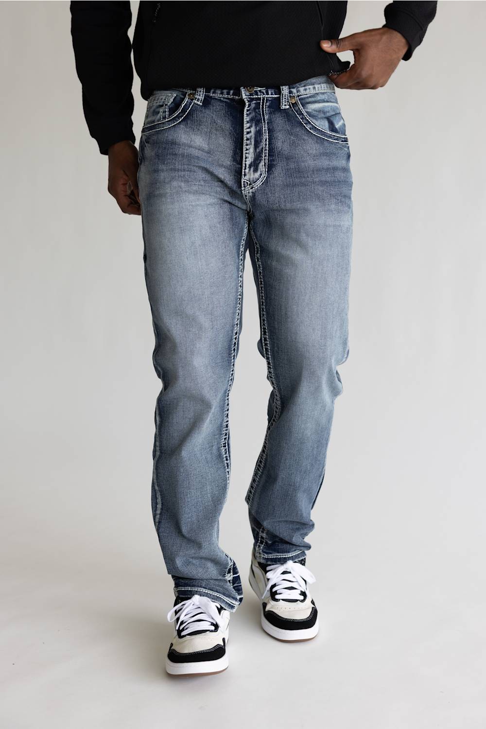 A person wearing light-wash blue denim jeans with a faded wash, paired with a black long-sleeve top and black-and-white sneakers, standing against a plain light gray studio background.