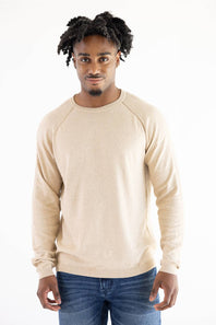 A man wearing a light beige knit raglan sweater and blue jeans, standing against a plain white background.