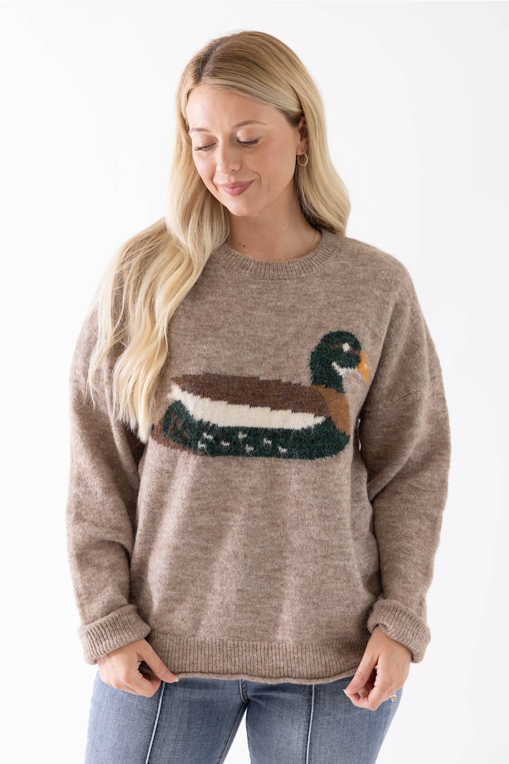 A woman wearing a taupe knit sweater with a large duck motif across the chest, paired with blue jeans, against a plain white background.