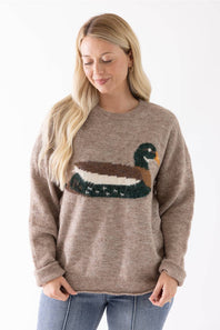 A woman wearing a taupe knit sweater with a large duck motif across the chest, paired with blue jeans, against a plain white background.