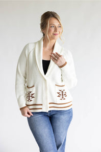 A woman modeling a cream-colored knit cardigan with brown geometric patterns on the pockets and hem, paired with blue jeans and a black top, against a plain light gray background.