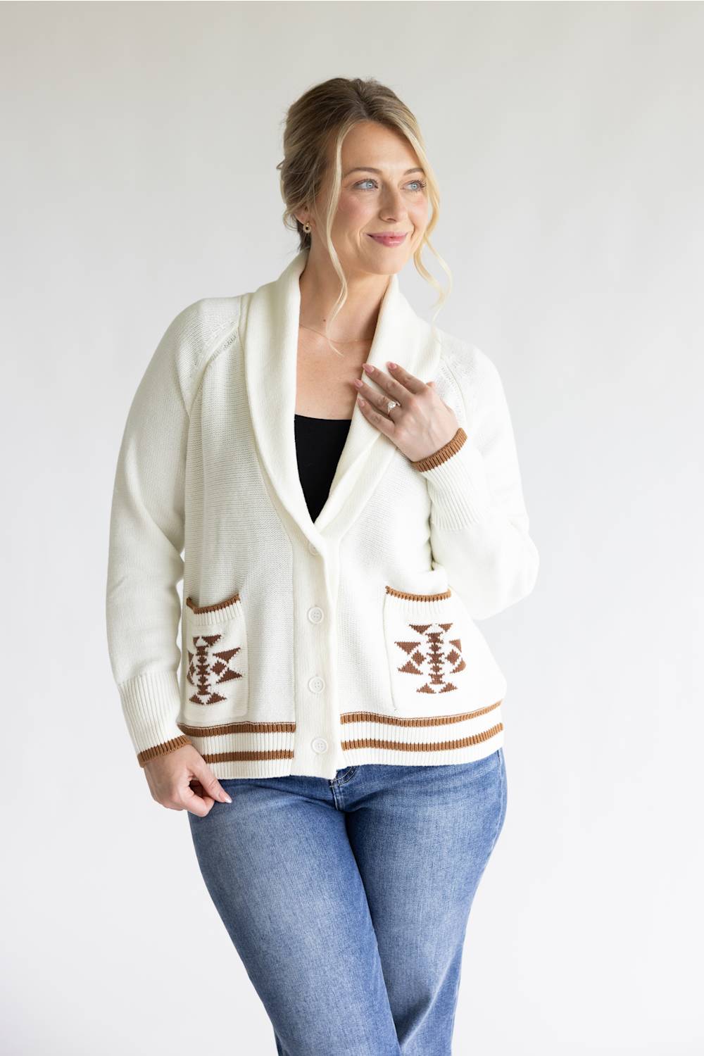 A woman modeling a cream-colored knit cardigan with brown geometric patterns on the pockets and hem, paired with blue jeans and a black top, against a plain light gray background.