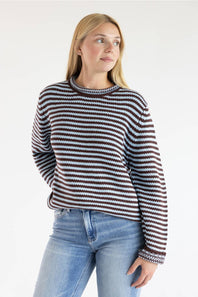 A woman wearing a long-sleeve knit sweater with horizontal brown, black, and light blue stripes, paired with light-wash blue jeans, against a plain white studio background.